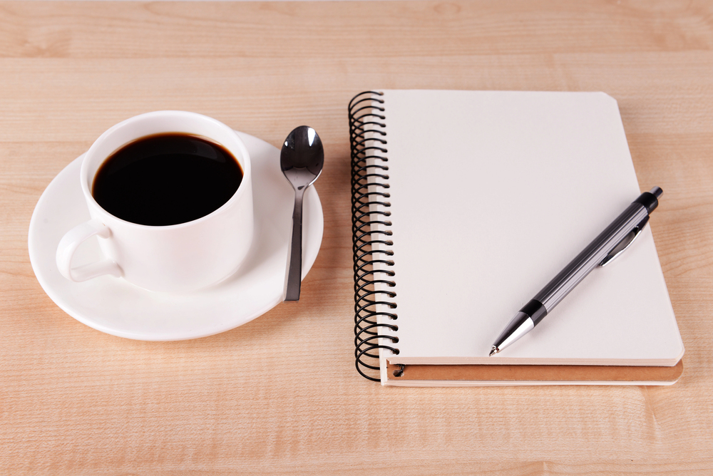 A white coffee cup on a white saucer with a spoon next to a spiral notebook and pen on a wooden surface